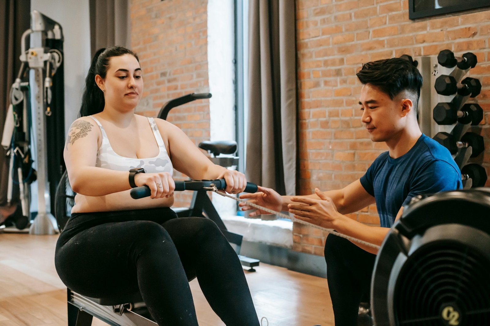 A personal trainer assists a woman on a rowing machine in a gym environment.